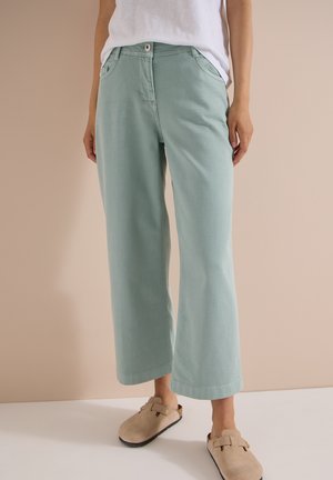 Personne portant un pantalon ample vert clair, un haut blanc et des chaussures beiges sans lacets, debout devant un fond beige uni.