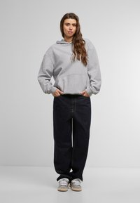 Felpa grigia con cappuccio e tasca frontale, abbinata a jeans blu scuri larghi. Sneakers bianche completano l'outfit casual.