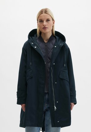 Femme portant une parka noire à capuche par-dessus une veste matelassée noire zippée et un jean bleu, debout devant un fond blanc uni.