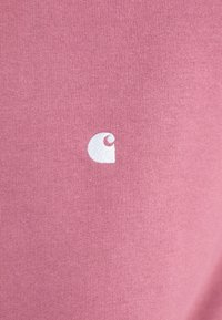 Rožinė medvilninė marškiniai su mažu baltu logotipu priekyje. Audinys atrodo švelnus, su lygiu paviršiumi ir klasikinio apykaklės dizainu.