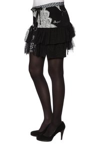 Schwarzer, gestuften Rock mit Spitzenbesatz und bestickten Details, ausgestattet mit einem gemusterten Taillenband und einer Schleife. Getragen über schwarzen Leggings und Absätzen.