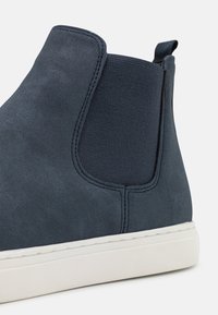 Námořnické kotníkové boty vyrobené z nubuku s elastickými bočními panely, hladkou texturou a bílou gumovou podrážkou. Žádné viditelné prvky ani vzory.