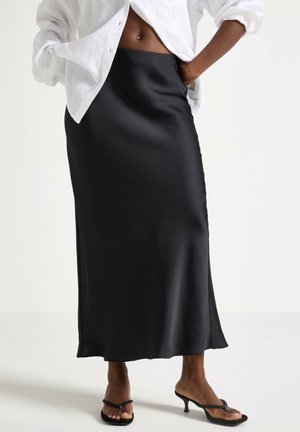 Person iført en sort satin midi nederdel, hvid skjorte med knapper og sorte sandaletter med hæl, stående med den ene hånd i lommen.