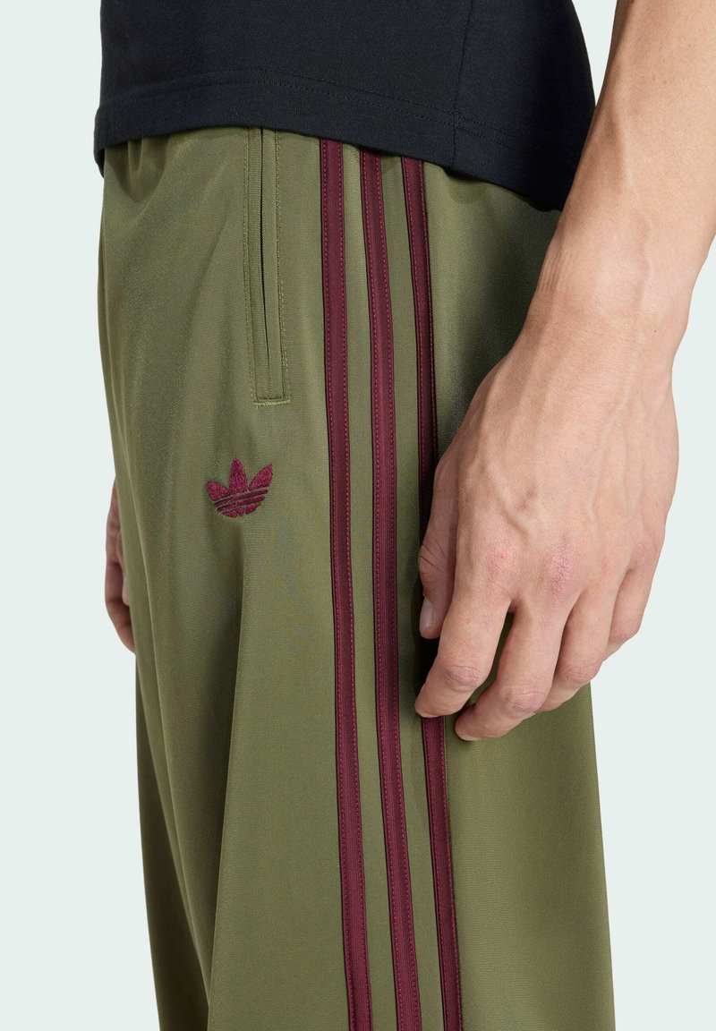 Olivgrüne Sporthose mit burgunderfarbenem Dreistreifen-Design, elastischem Bund und gesticktem Logo am linken Oberschenkel. Glatte Textur.