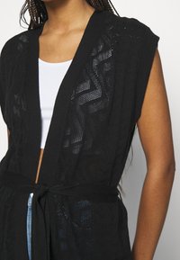 Chaqueta kimono negra de punto con mangas cortas, que presenta un patrón de recortes geométricos y un cierre de lazo en la cintura. Textura ligera.