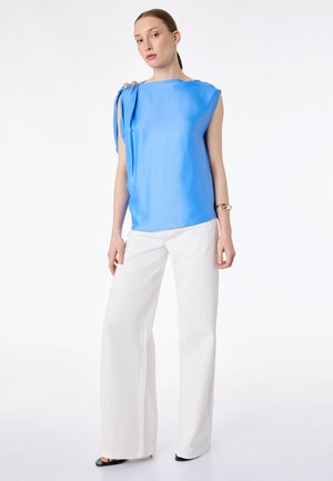 Kobieta w bezrękawniku w kolorze błękitnego nieba z detalem zawiązanym na ramieniu oraz białych, szerokich spodniach, stojąca na tle białego tła.