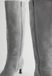 Botas altas de ante gris con una silueta slim y un tacón bajo ancho. Cuentan con una cremallera lateral para facilitar su uso y una textura suave.
