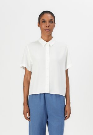 Kurzes, weißes Blusenshirt mit spitzem Kragen, Knopfverschluss und lässiger Passform. Getragen mit blauen Hosen. Glatte Textur.