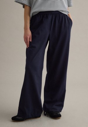 Persona con pantalones sueltos azul marino con cintura elástica, zapatillas negras y una camisa de manga corta a rayas, de pie con las manos en los bolsillos.