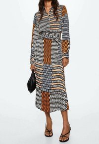 Femme portant une robe midi à manches longues avec des motifs géométriques orange, noir et blanc, tenant un sac noir et chaussée de sandales noires à talons.