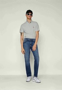 Uomo vestito con polo grigia, jeans blu, sneakers bianche, berretto nero e occhiali da sole, in piedi contro uno sfondo semplice.