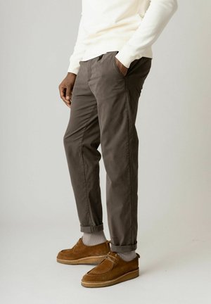 Oseba, oblečena v rjave zavihane hlače, rjave semiš čevlje, bele nogavice in belo dolg rokav, z eno roko v žepu.