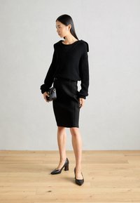 Pull noir à côtes avec un large décolleté, associé à une jupe noire ajustée avec des boutons. Des talons noirs complètent l'ensemble, posé sur un sol en bois.