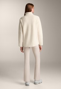 Pullover in pile color crema, morbido e soffice, con colletto alto, abbinato a pantaloni beige attillati e scarpe grigio chiaro, visto da dietro.