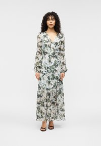 Μακρύ, λουλουδάτο φόρεμα από ελαφρύ ύφασμα με πράσινα και κρεμ λουλουδένια σχέδια. Σχεδίαση τύπου wrap με μακριά μανίκια και φούστα με βολάν, σε συνδυασμό με μαύρα σανδάλια.