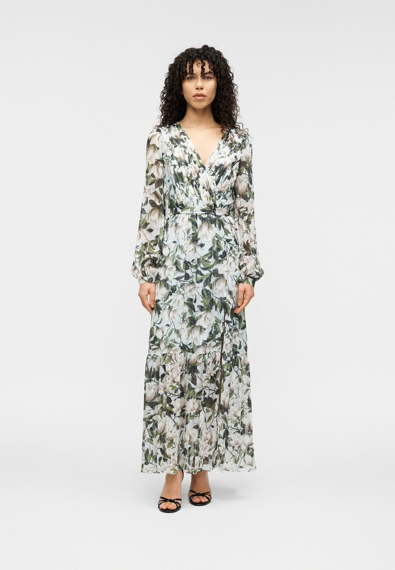 Μακρύ, λουλουδάτο φόρεμα από ελαφρύ ύφασμα με πράσινα και κρεμ λουλουδένια σχέδια. Σχεδίαση τύπου wrap με μακριά μανίκια και φούστα με βολάν, σε συνδυασμό με μαύρα σανδάλια.