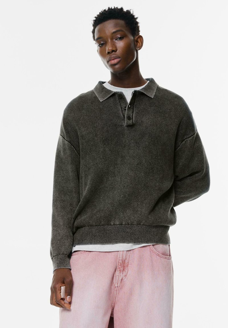 Donkergroene trui met een kraag, knoopdetail aan de voorkant, oversized pasvorm, ribgebreide zoom en verlaagde schouders, gecombineerd met lichtroze broek.