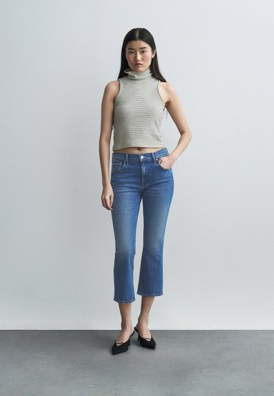 Γυναίκα που στέκεται μπροστά σε απλό φόντο, φορώντας αμάνικο ριγέ τοπ με ζιβάγκο, κοντά τζιν και μαύρες μυτερές γόβες.