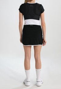 Černý sportovní outfit zahrnující přiléhavý top s síťovaným designem a černou sukni s bílým lemem, doplněný bílými ponožkami a botami.