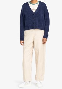 Marinblå kort kofta med V-ringning, fem svarta knappar, ribbstickade muddar och en mjuk textur, kombinerad med beige vida byxor och vita sneakers.