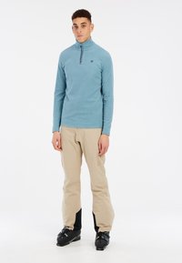 Pullover in pile azzurro chiaro con colletto a zip, abbinato a pantaloni da sci beige e scarponi da sci neri. Il pullover presenta un logo discreto sul petto.