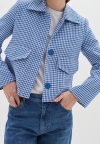 Chaqueta de cuadros azul y blanca con cuello, dos bolsillos frontales y botones azules, combinada con jeans de mezclilla azul. Textura suave.