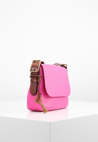 Bolso cruzado de cuero rosa con cinturón ajustable marrón y un charm de llave dorado colgando del broche del cinturón, exhibido sobre una superficie blanca.