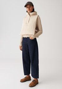 Beige Daunenweste mit hohem Kragen, kombiniert mit einem gerippten cremefarbenen Top und weiten, marineblauen Hosen. Braune Schnürschuhe im minimalistischen Design.