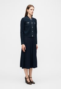 Γυναίκα που φοράει μια ναυτική ζακέτα με λευκές ραφές, μια αντίστοιχη πλισέ midi φούστα και καφέ ψηλοτάκουνα παπούτσια με λουράκι Τ, στέκεται μπροστά σε λευκό φόντο.