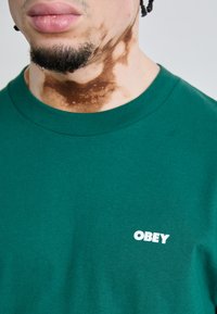 Zöld pamut póló, kerek nyakkivágással. A bal mellen fehér "OBEY" logó található. Az anyaga sima és nem textúrázott.