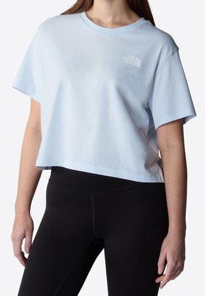 Kobieta w jasnoniebieskim krótkim T-shircie z logo The North Face oraz czarnych legginsach, stojąca na białym tle.