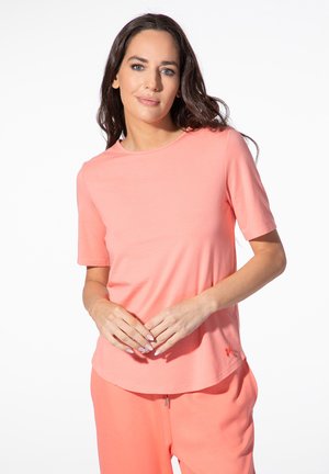 Woman with long dark hair wearing a coral short-sleeve shirt and matching pants, standing with hands gently clasped.