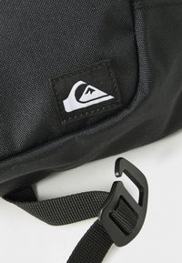 Black textured fabric bag with a white logo patch and an adjustable black strap featuring a plastic hook buckle on a white surface.