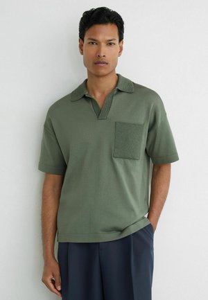 Mann steht an weißer Wand, trägt olivgrünes kurzärmeliges Poloshirt mit Brusttasche und marineblaue Hose, Hände entspannt.