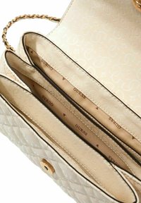 Sac à main beige matelassé avec une fermeture à bouton doré et une bandoulière chaîne. L'intérieur comprend trois compartiments, doublés de tissu avec le logo "GUESS".