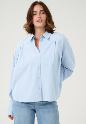 Himmelblaues Button-Up-Hemd mit lässiger Passform, das einen Kragen, gefaltete Schultern und lange Ärmel bietet. Getragen mit heller Jeans.
