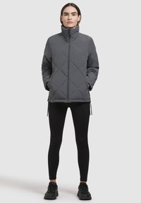 Gray quilted puffer jacket with high collar, front zipper, and side pockets. Worn over black leggings and sneakers.