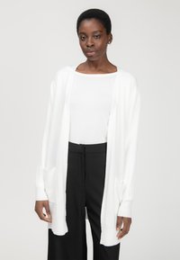 Witte gebreide cardigan met open voorkant, lange mouwen en twee voorkwabben, gecombineerd met een witte top en zwarte high-waisted broek. Gladde textuur.