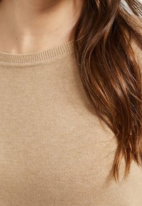 Beige knitted sweater with a ribbed neckline, featuring a smooth texture and close-fitting design, partially obscured by long, wavy hair.