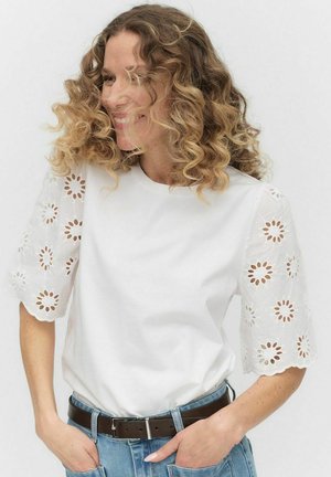 Hvid bomulds t-shirt med broderede blondeærmer, der har blomstermønstre. Båret med lyseblå denim og et brunt bælte.