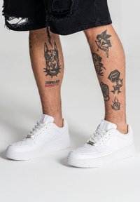 Sneaker bianche con un motivo textured, suola in gomma spessa e design con lacci. I dettagli includono un logo metallico e cuciture intricate.