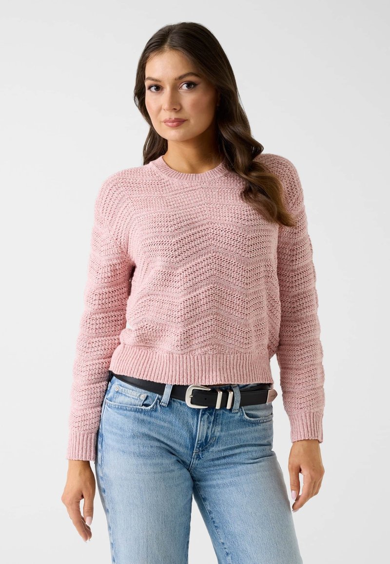 Pull tricoté rose clair avec une texture ondulée, col rond et ourlet côtelé, associé à un jean bleu clair et une ceinture noire.