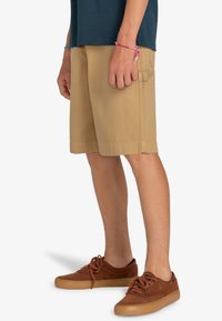 Shorts beiges en coton robuste, dotés de poches latérales et d'une coupe droite. Associés à des baskets en daim marron et une chemise bleu foncé.
