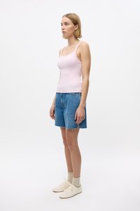 Gaiši rozā bez piedurknēm topiņš, džinsu šorti ar saplaisājušām malām un baltas kedas, kas apvienotas ar bēša zeķēm. Modelis stāv profila pozīcijā pret vienkāršu fona.