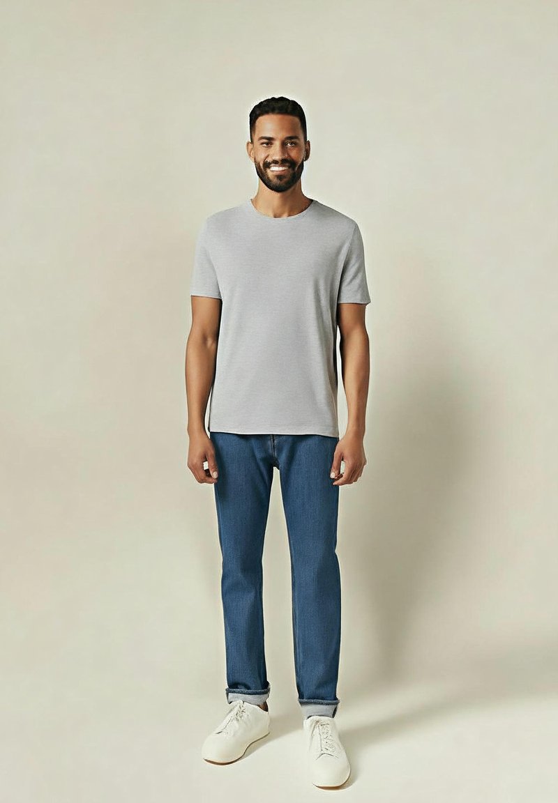 Hombre de pie y sonriendo, vestido con una camiseta de manga corta gris claro, jeans azul doblados y zapatillas blancas sobre un fondo plano.