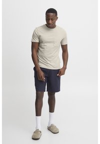 Tricou din bumbac bej deschis cu un buzunar pe piept, asociat cu şorturi negre închis. Accesoriile includ şosete albe şi pantofi slip-on bej.