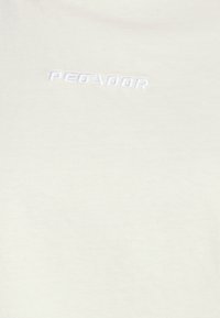 Vaaleanbeige t-paita, joka on valmistettu pehmeästä puuvillasta ja jossa on kohotettu valkoinen brodeerattu logo "PEGADOR" keskellä rintaa.