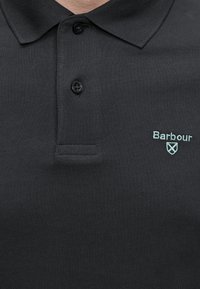 Polo noir avec deux boutons noirs et logo Barbour bleu clair ainsi qu'un emblème en forme de bouclier sur la poitrine.