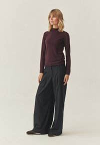 Burgundská košile s dlouhým rukávem v kombinaci s širokými kalhotami s námořnickými proužky, které mají hladkou texturu a volný střih. Outfit doplňují hnědé boty.