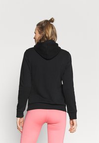 Felpa nera con cappuccio, caratterizzata da maniche lunghe e orlo a coste. Chi la indossa la abbina a legging rosa. La vista posteriore mostra un design minimalista.